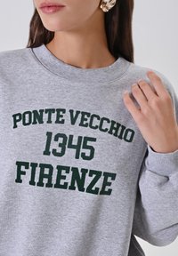 Donna che indossa una felpa grigia con testo verde scuro "Ponte Vecchio 1345 Firenze" e grandi orecchini d'argento, mano che tocca il colletto.