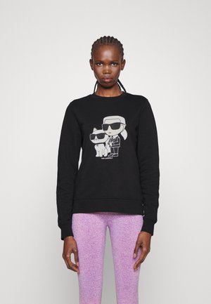 Sudadera de algodón negra con un gráfico de dos personajes ilustrados que llevan gafas de sol. Puños y dobladillo acanalados, diseño minimalista.