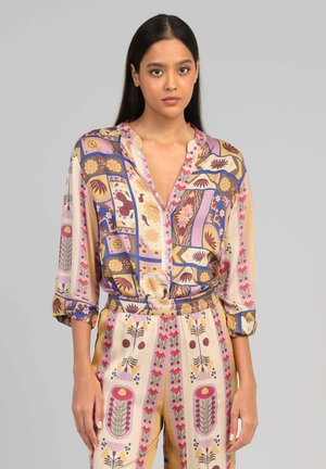 Femme aux longs cheveux foncés portant un chemisier coloré à motifs assorti à un pantalon taille haute avec des motifs floraux et abstraits.