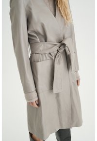 Kobieta ubrana w wiązany jasnoszary skórzany płaszcz trench z dużymi przednimi kieszeniami, pod którym ma beżowy sweter z długim rękawem oraz ciemne spodnie.