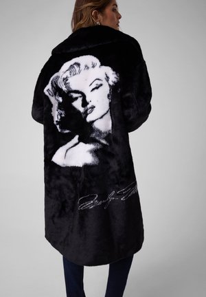 Une femme porte un long manteau noir orné d’un grand portrait blanc de Marilyn Monroe et d’une signature dans le dos.