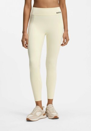 Kvinde iført lysegule højttaljede leggings med BOSS-logo og beige sneakers, stående mod en hvid baggrund.