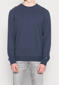 Tmavě modrá mikina s dlouhými rukávy a kulatým výstřihem, s žebrovanými manžetami a lemem, zdobená malým detailem loga na levém zápěstí.