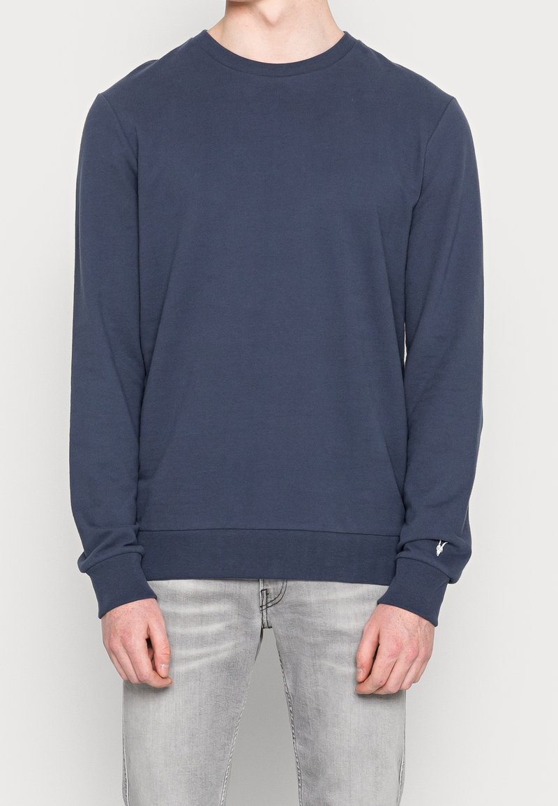 Tmavě modrá mikina s dlouhými rukávy a kulatým výstřihem, s žebrovanými manžetami a lemem, zdobená malým detailem loga na levém zápěstí.