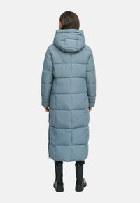 Woman wearing a long, light blue quilted puffer coat with a hood, black pants, and black ankle boots, standing facing away on a white background.