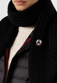 Persona vistiendo una bufanda de punto grueso negra, una chaqueta acolchada negra con ribetes rojos y un suéter de punto blanco, mostrando un pequeño parche de logo circular.