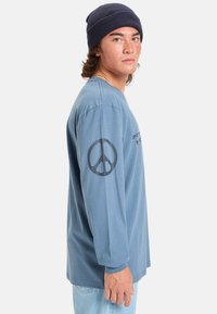 Camiseta de manga larga azul, de material de algodón, con un símbolo de paz en la manga izquierda y texto negro en el pecho. Lleva puesta una gorra de lana azul marino.
