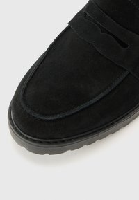 Zwarte suede moccasin met een platte ontwerp, voorzien van een decoratieve uitsnijding en een duurzame zwarte rubberen zool.