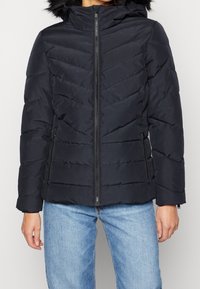 Marineblauer Puffer-Jacke mit Reißverschluss, Chevron-Steppung und einer mit Kunstpelz gefütterten Kapuze. Verfügt über Seitentaschen und elastische Bündchen.