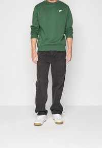 Grön Nike-tröja med rundhals, kombinerad med mörkgrå jeans. Vita sneakers har en beige detalj på sulan. Enkla bakgrund.