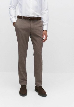 Bruine op maat gemaakte broek van een zachte, gestructureerde stof. Voorzien van een knoop- en ritssluiting, met een bruine riem en bruine schoenen.