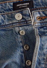 Jeans in denim blu con accessori in ottone, dotati di chiusura a bottoni e cuciture arancioni. L'etichetta recita "JACK & JONES" in vita.