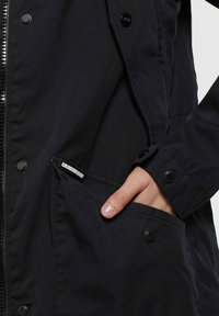 Svart jacka med dragkedja, tryckknappar och en sidoficka. Materialet är slätt med en matt yta. Innehåller en logotagg nära fickan.