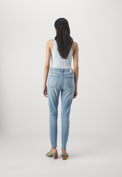 Fakó kék, slim fit farmernadrág magas derékkal, klasszikus ötfakkos dizájnnal és rojtos szegéllyel. Zöld magassarkú szandálokkal viselve.