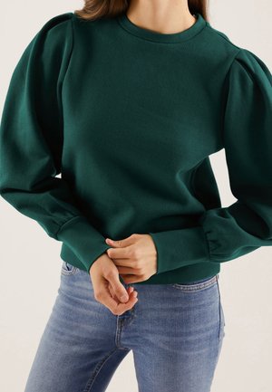 Frau trägt dunkelgrünes Top mit Puffärmeln und hellblaue Jeans, Hände am Taillenbereich verschränkt, vor schlichtem Hintergrund.