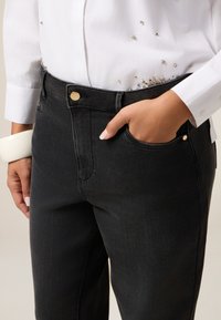 Los pantalones de mezclilla negros cuentan con un corte clásico, bolsillo delantero y un acento de botón dorado. La tela tiene una textura suave y un desvanecimiento mínimo.