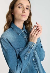 Woman with light skin and long brown hair wearing a blue denim shirt, holding her hands together near her face, neutral expression.