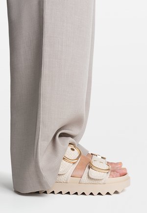 Sandales à plateforme beiges avec des sangles tissées texturées et des accessoires en or, associées à un pantalon gris clair, à jambe large et en tissu doux.