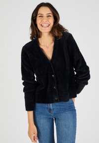 Cardigan nero e soffice con scollo a V, maniche lunghe e chiusura con cinque bottoni, abbinato a jeans blu. Tessuto morbido e vestibilità rilassata.