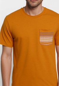 Camiseta de rayas naranja con bolsillo a rayas en tonos marrón y crema. Mangas cortas, escote redondo y corte relajado.