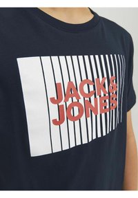 Jūras spēku kokvilnas T-krekls ar baltu taisnstūra grafiku, kurā ir vertikālas līnijas un zīmola nosaukums "JACK & JONES" sarkanos burtos.