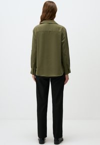 Olijfgroene shirt met een ontspannen pasvorm, lange mouwen, knoopmanchetten en een rechte zoom, gecombineerd met zwarte zakelijke trousers en zwarte schoenen.