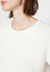 Pull en maille couleur crème avec une texture côtelée, un col rond et des manches raglan, présenté dans un tissu doux et léger.