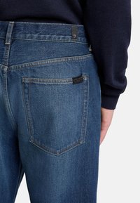 Niebieskie dżinsy o prostym kroju, wyraźnym przeszyciu oraz czarnej metce na pasku, z detalem kieszeni z tyłu.