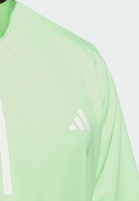 Maglietta sportiva verde chiaro con colletto alto. Presenta una cerniera verticale bianca e un piccolo logo bianco con tre strisce sul petto sinistro.