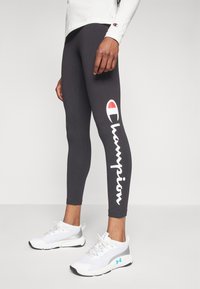 Svarta leggings med vit "Champion"-logotyp på högra benet, tillsammans med vita träningssneakers med ljusblå accenter och grå detaljer.