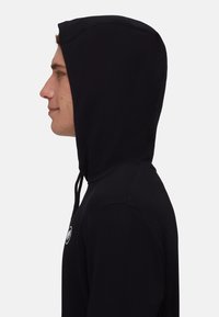 Haut à capuche noir avec cordon de serrage, vue de côté montrant la texture douce du tissu et un logo rond sur la poitrine. Design simple et ajusté.