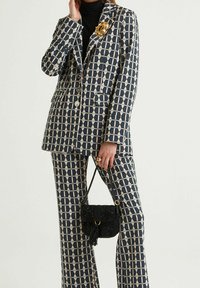 Costume à motifs bleu marine et crème composé d'un blazer cintré et d'un pantalon large. Comprend une broche dorée et un petit sac noir à sequins.