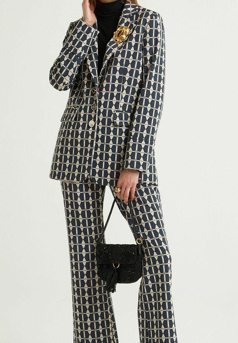 Costume à motifs bleu marine et crème composé d'un blazer cintré et d'un pantalon large. Comprend une broche dorée et un petit sac noir à sequins.