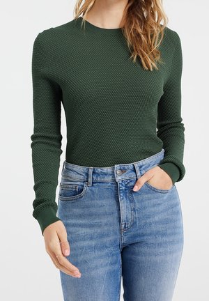 Mujer con suéter de manga larga texturizado verde oscuro metido dentro de unos vaqueros de tiro alto azul claro, con una mano en el bolsillo.