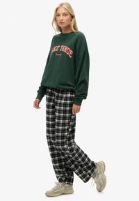 Grüner Sweatshirt mit der Aufschrift „LAKE TAHOE“ in pinken Buchstaben, kombiniert mit schwarz-weiß karierten Hosen und hellen Sneakers.