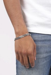 Silver metal chain bracelet worn on the wrist, featuring interlocking links and a polished finish, paired with a white shirt and blue jeans.