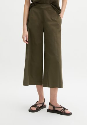 Femme portant un pantalon large court vert olive et des sandales plates noires à brides, debout les mains dans les poches.