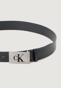 Curea din piele neagră, cu o cataramă metalică rectangulară argintie cu logo-ul tăiat "CK". Curea are o textură netedă și o ajustare variabilă.