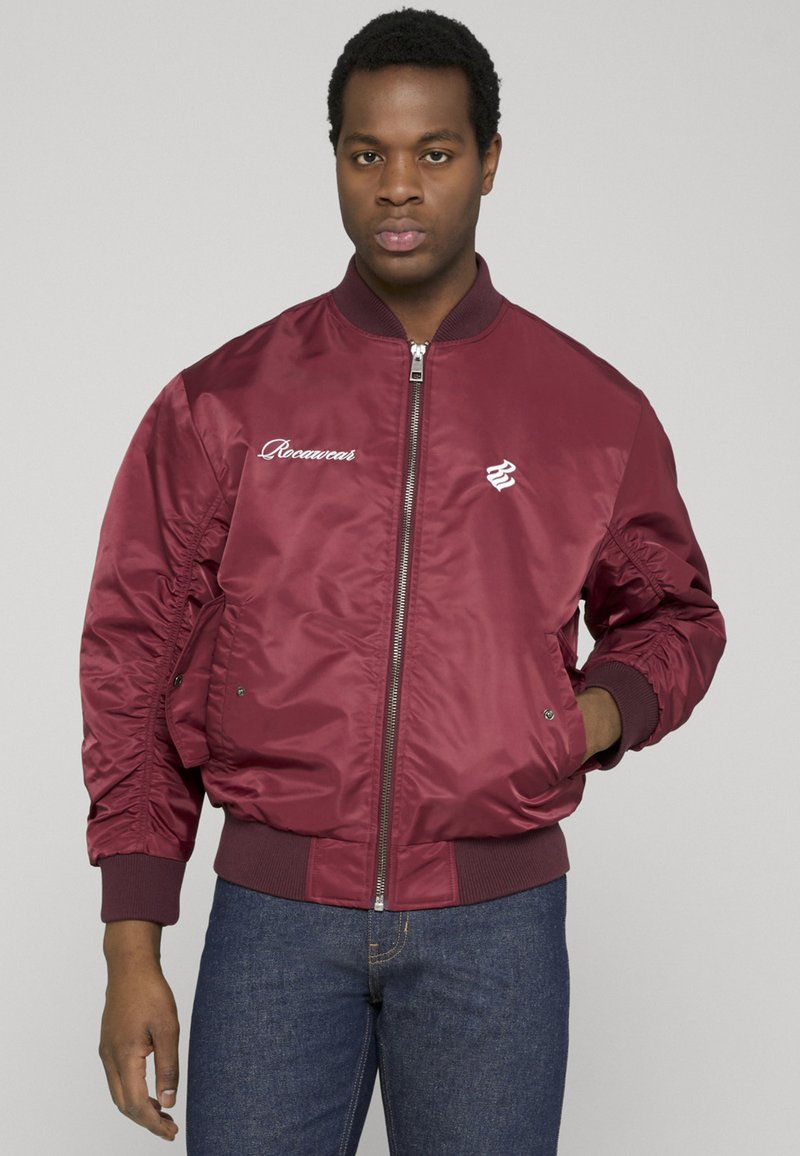 Homme portant une veste bomber bordeaux zippée avec des logos sur la poitrine et un jean bleu, debout devant un fond clair uni.