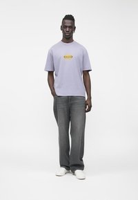 Fialové tričko s krátkym rukávom a logom 'GUESS', skombinované so sivými džínsami s širokými nohavicami a bielymi teniskami, na jednoduchom pozadí.