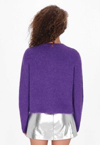 Lila, bolyhos kötött pulóver hosszú ujjakkal és kerek nyakkivágással, tükröződő ezüst mini szoknyával párosítva. Hátulnézet látható.