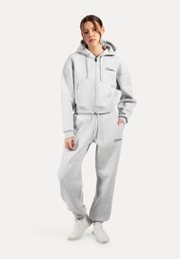 Felpa grigia con zip e tasca frontale, abbinata a pantaloni della tuta grigi coordinati. Entrambi presentano una texture morbida e accenti neri eleganti.