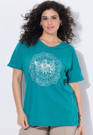 Kobieta z kręconymi włosami ubrana w morski t-shirt z białym motylem i kwiatowym wzorem mandali, stojąca na białym tle.