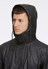 Svart vattentät jacka med hög krage och justerbar snoddhuva. Slät textur med tryckknapps-stängningar och minimalistisk design.
