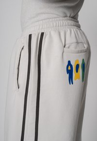 Graue Jogginghose aus Baumwolle mit seitlichen Taschen, schwarzen Streifen und einem bunten Motiv von Figuren in Gelb, Grün und Blau am Oberschenkel.