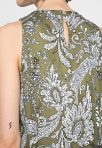 Blusa senza maniche verde oliva con un motivo floreale bianco, schienale con taglio a chiave e chiusura a bottone, e tessuto texturizzato.