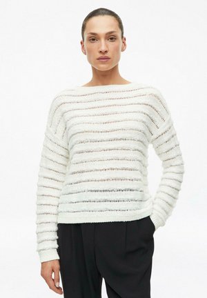 Vrouw met naar achteren getrokken haar, gekleed in een witte gestreepte gebreide trui en zwarte broek, staand met één hand in de zak tegen een witte achtergrond.