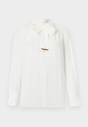 Witte blouse met lange mouwen, geplooide schouders en een grote strik bij de kraag, met gouden manchetknoopjes.