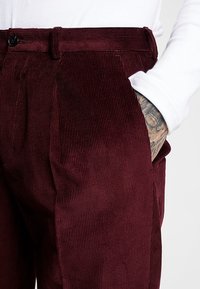 Burgundy kordfluwelbyxor med vertikal ribbning, som har en framficka och knäppe. Inga synliga mönster; slät textur.