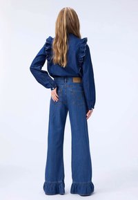 Denim topp med volanger på skuldrene og lange ermer, kombinert med vide bukser med frynser nederst og lommer bak.